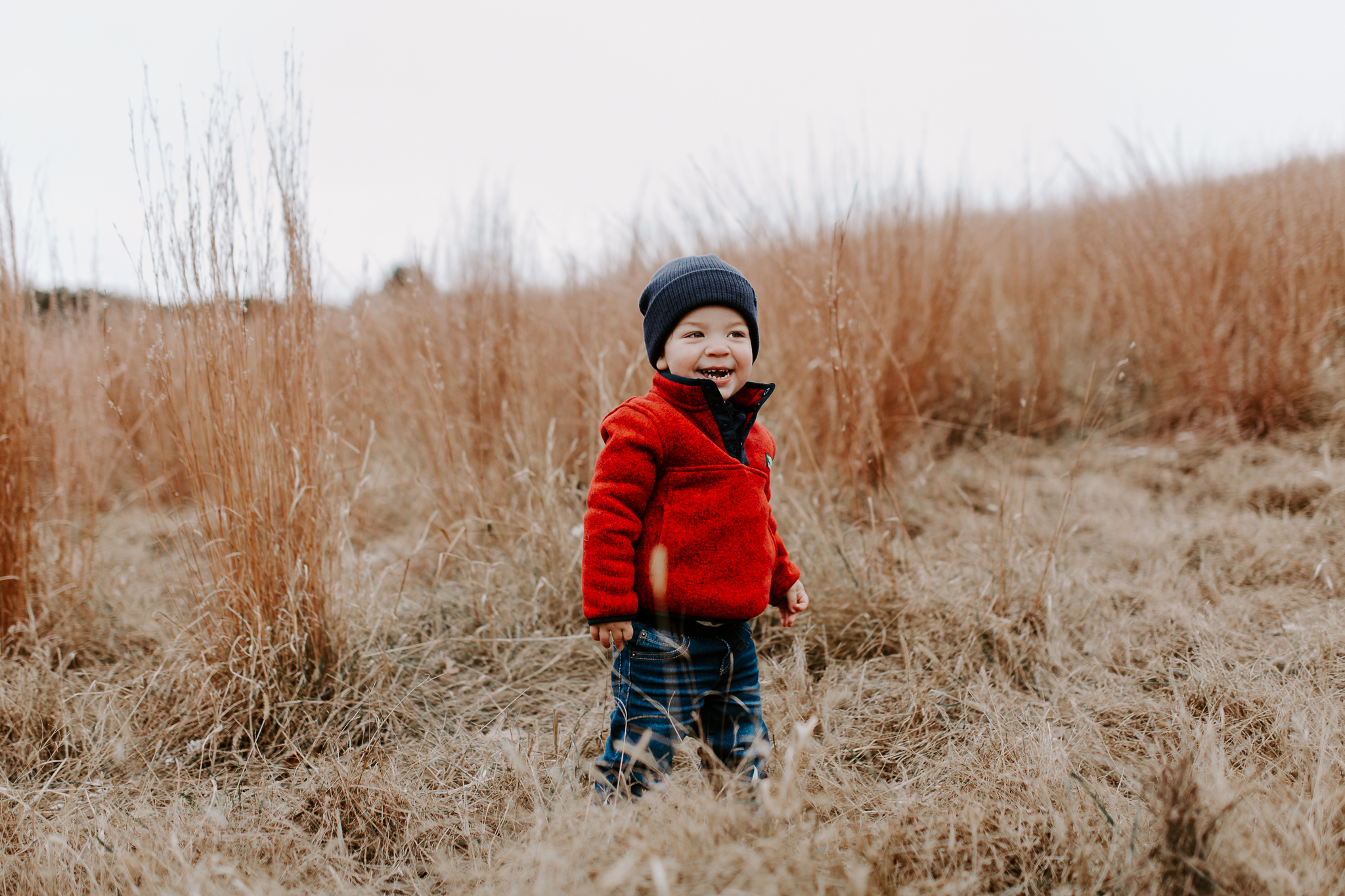 Young boy in a red coat, smiling alone in a fall field of dried grasses.
